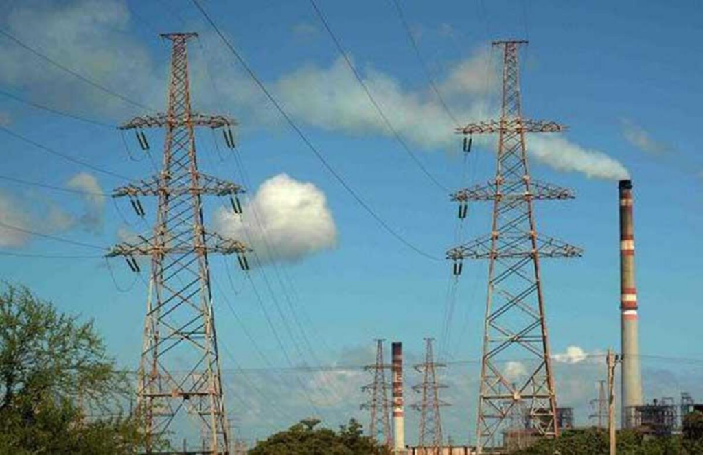 Torres de alta tensión y líneas eléctricas con una planta de energía y chimeneas industriales al fondo bajo un cielo azul.