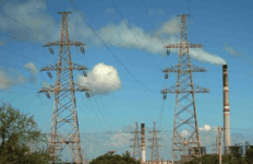Torres de alta tensión y líneas eléctricas con una planta de energía y chimeneas industriales al fondo bajo un cielo azul.