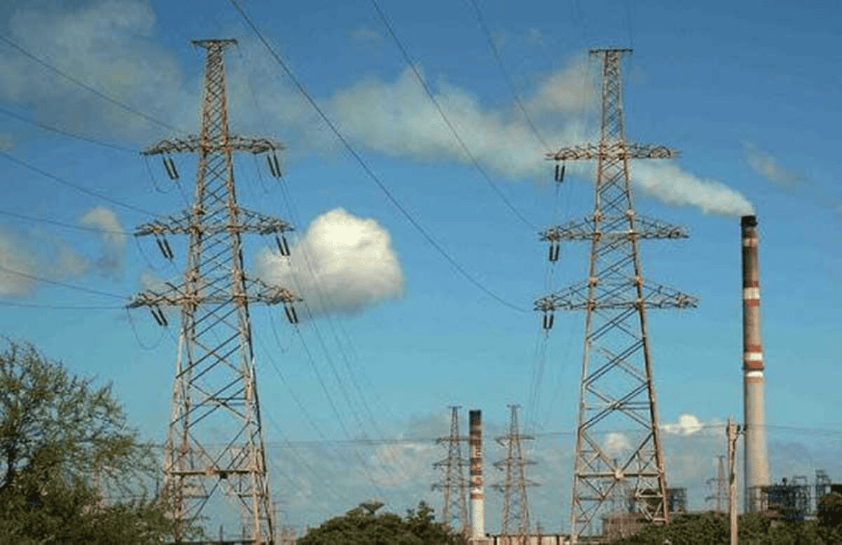 Torres de alta tensión y líneas eléctricas con una planta de energía y chimeneas industriales al fondo bajo un cielo azul.