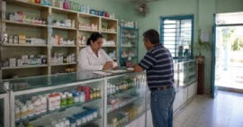 Interior de una farmacia inspirada en Cuba con estanterías de medicamentos, mostrador de atención y un farmacéutico atendiendo a un cliente.