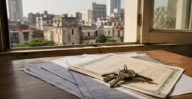 Llaves de una vivienda sobre documentos de propiedad en el interior de una casa cubana, con edificios residenciales desenfocados al fondo, como representación de la nueva Ley de la Vivienda en Cuba.