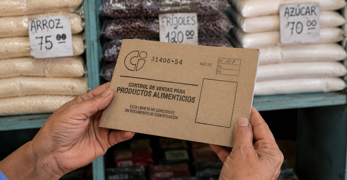Manos sosteniendo una libreta de abastecimiento en una bodega cubana con arroz, frijoles y azúcar al fondo.