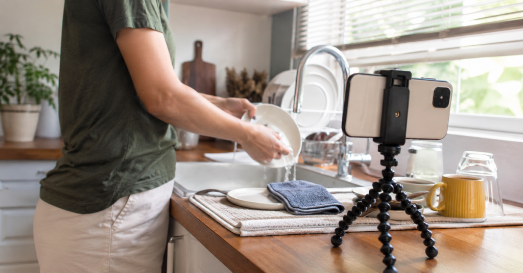 Persona grabándose con un teléfono móvil mientras lava platos en una cocina para participar en tareas de entrenamiento de inteligencia artificial.