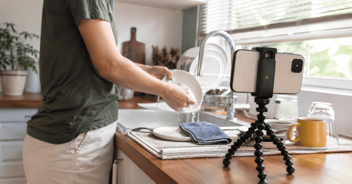Persona grabándose con un teléfono móvil mientras lava platos en una cocina para participar en tareas de entrenamiento de inteligencia artificial.