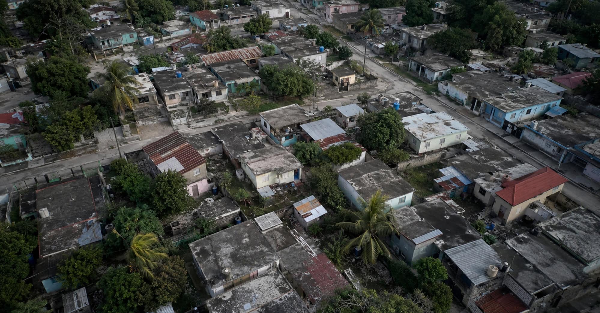 Vista aérea ilustrativa de un barrio cubano con viviendas modestas, techos desgastados y vegetación tropical.