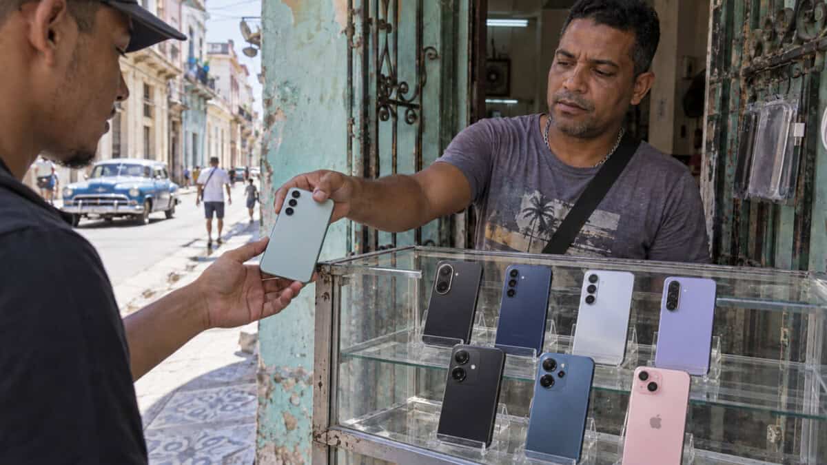 Vendedor entrega un teléfono a un cliente junto a una vitrina con celulares en un puesto de ambiente cubano.