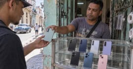 Vendedor entrega un teléfono a un cliente junto a una vitrina con celulares en un puesto de ambiente cubano.
