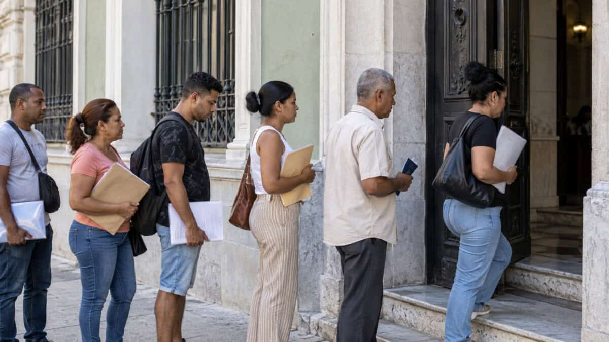 Ciudadanos cubanos con carpetas y documentos esperan en fila frente a un edificio de arquitectura habanera.