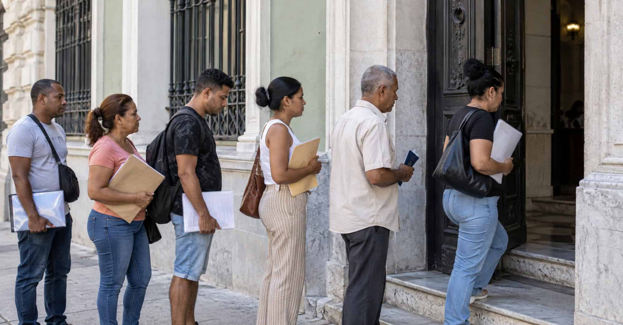 Ciudadanos cubanos con carpetas y documentos esperan en fila frente a un edificio de arquitectura habanera.