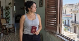 Mujer en una vivienda cubana sostiene un pasaporte español mientras mira por una ventana.