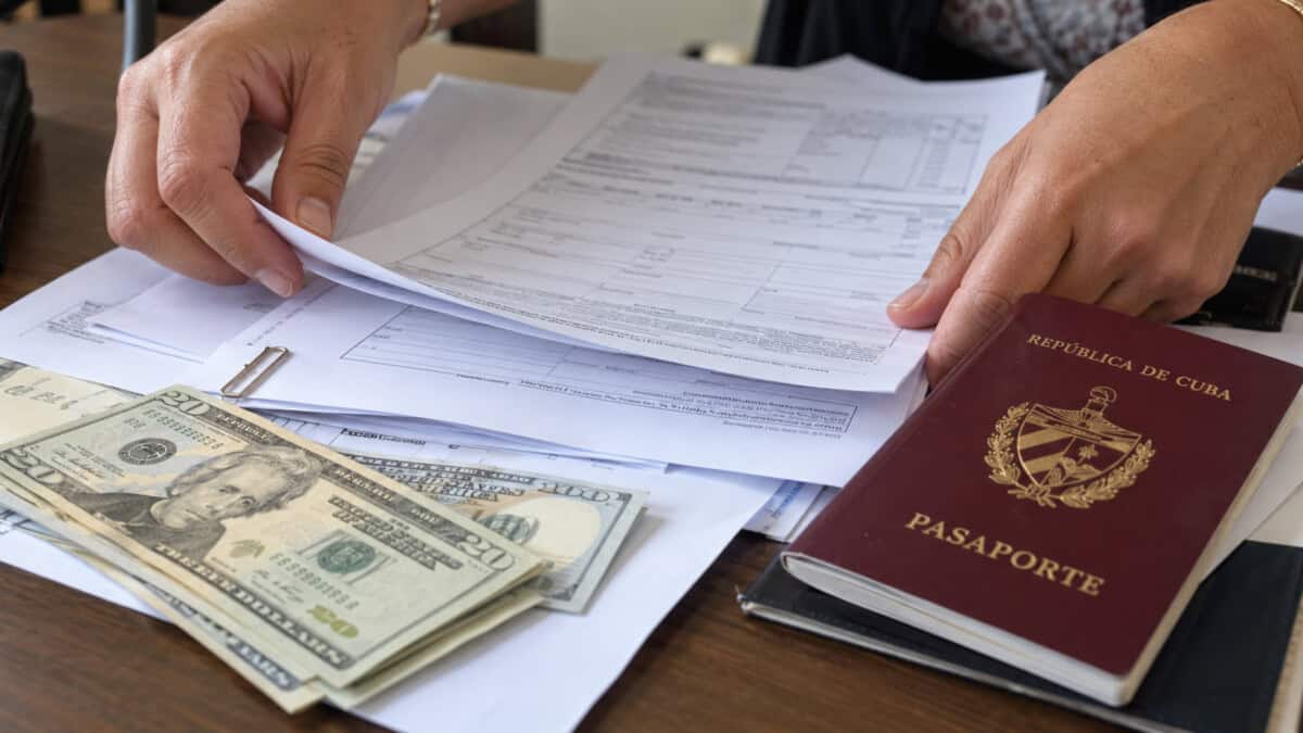 Manos organizando formularios junto a un pasaporte cubano y billetes de dólares estadounidenses.
