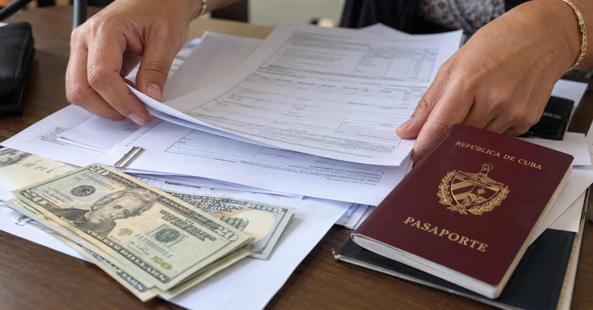 Manos organizando formularios junto a un pasaporte cubano y billetes de dólares estadounidenses.
