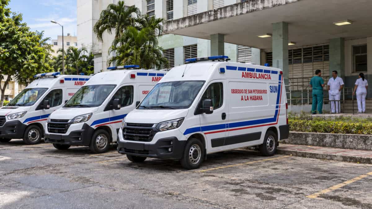 Tres ambulancias blancas con franjas rojas y azules frente a un hospital, en una imagen conceptual del sistema sanitario cubano.