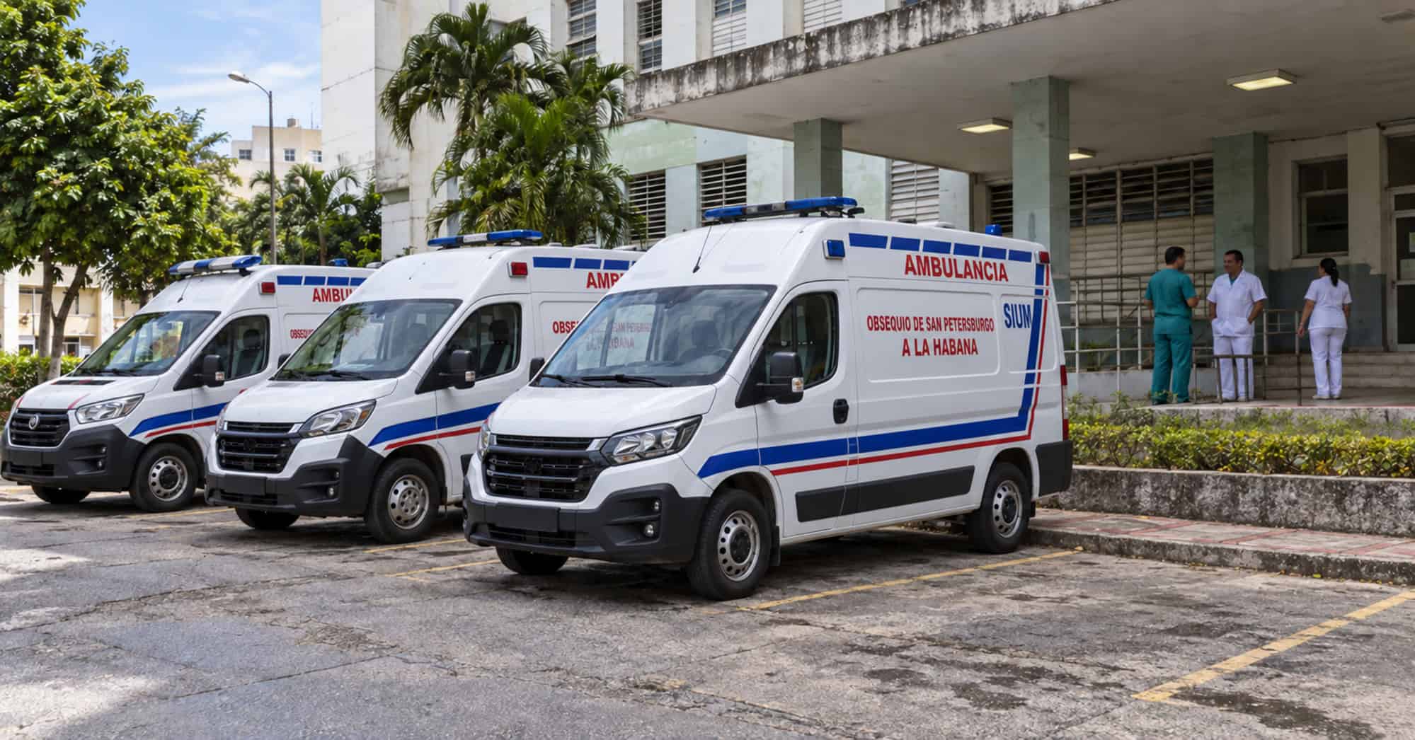 Tres ambulancias blancas con franjas rojas y azules frente a un hospital, en una imagen conceptual del sistema sanitario cubano.