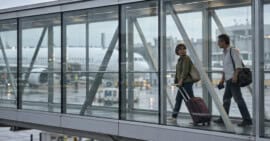 Pareja con equipaje y un pasaporte cerrado camina por un pasillo acristalado de un aeropuerto, con un avión al fondo.