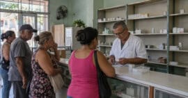 Farmacéutico atiende a varios clientes en una farmacia comunitaria cubana con estanterías parcialmente abastecidas.