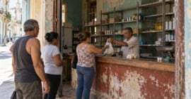 Compradores frente al mostrador de un comercio de barrio cubano con alimentos y productos básicos en estantes parcialmente vacíos.