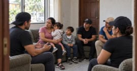 Grupo de adultos y dos niños sentados conversando en la sala de un hospedaje, en una recreación ambientada en Tapachula, México.
