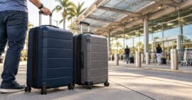 Viajero con dos maletas cerradas frente a la entrada de un aeropuerto, con dos personas desenfocadas al fondo.