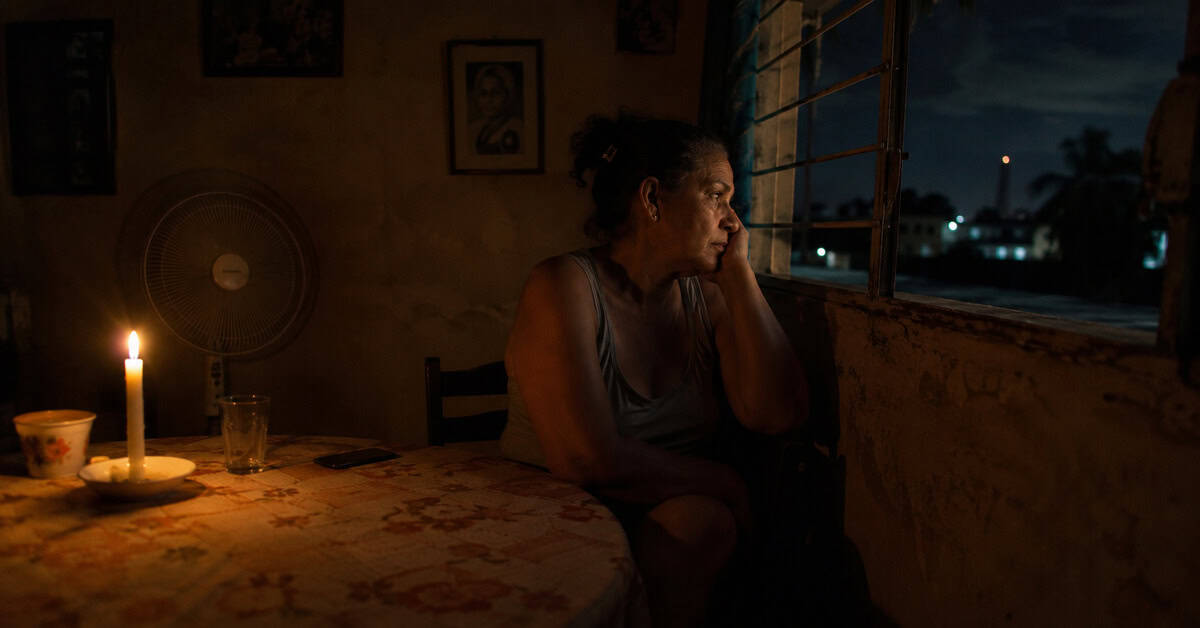 Interior de una vivienda cubana durante un apagón nocturno con una persona sentada junto a una ventana iluminada únicamente por la luz de una vela.