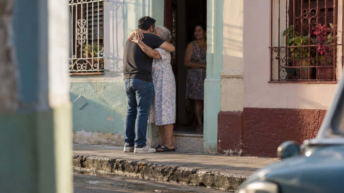 Un hombre abraza a una mujer mayor frente a una vivienda cubana, mientras otra mujer observa desde la puerta.