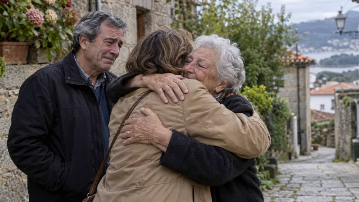 Mujer mayor abraza a una familiar junto a un hombre en una calle de piedra de una localidad gallega.