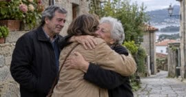 Mujer mayor abraza a una familiar junto a un hombre en una calle de piedra de una localidad gallega.