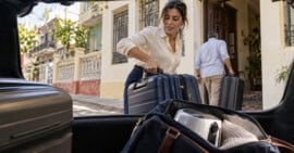 Mujer retira una maleta del maletero de un automóvil mientras otro viajero lleva su equipaje hacia una vivienda cubana.