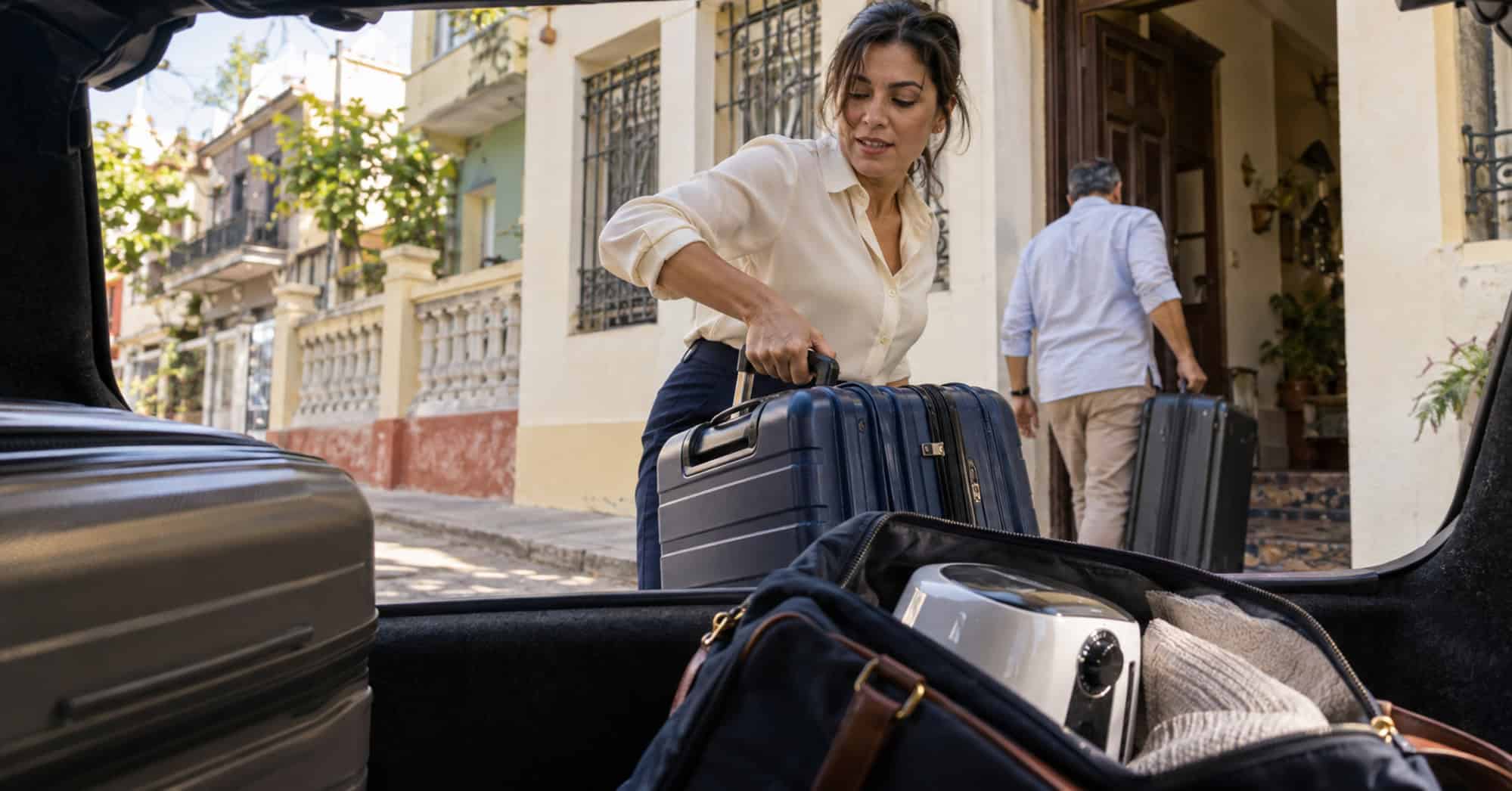 Mujer retira una maleta del maletero de un automóvil mientras otro viajero lleva su equipaje hacia una vivienda cubana.