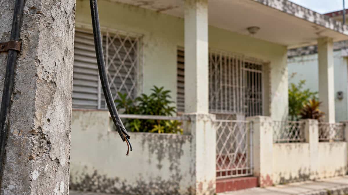 Cable cortado colgando junto a un poste, con una vivienda cubana desenfocada al fondo.