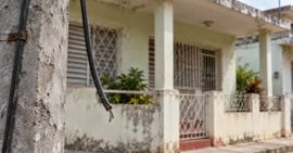 Cable cortado colgando junto a un poste, con una vivienda cubana desenfocada al fondo.