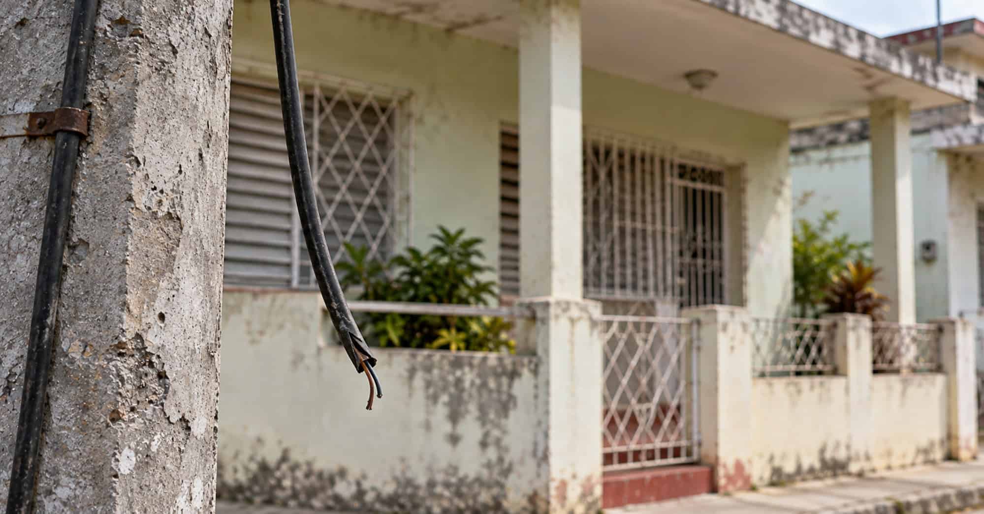 Cable cortado colgando junto a un poste, con una vivienda cubana desenfocada al fondo.