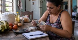 Mujer cubana cuenta billetes en una mesa con calculadora, libreta y alimentos básicos.