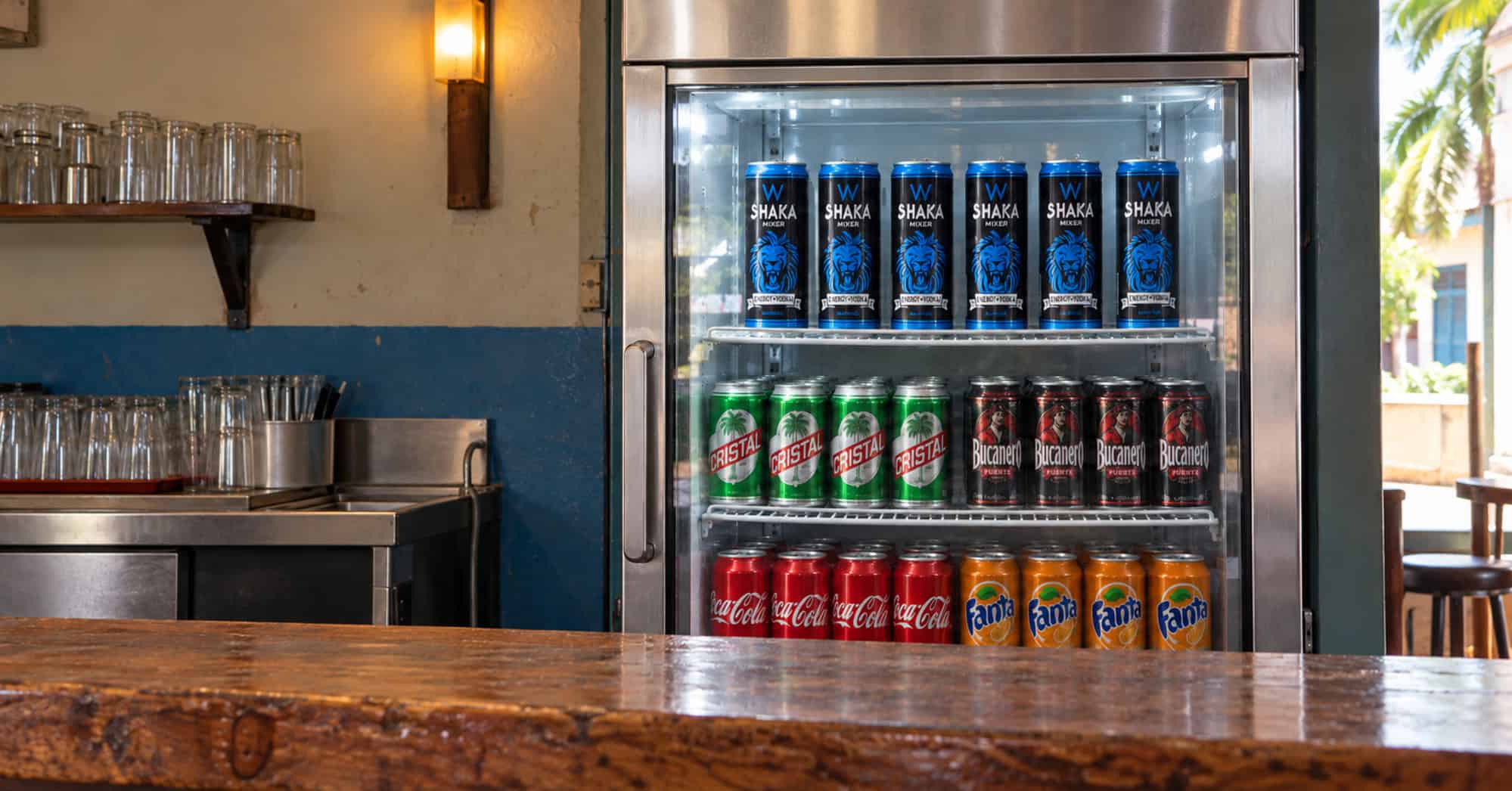 Seis latas de Shaka en un exhibidor con puerta de cristal, junto a cervezas Cristal, Bucanero y refrescos.