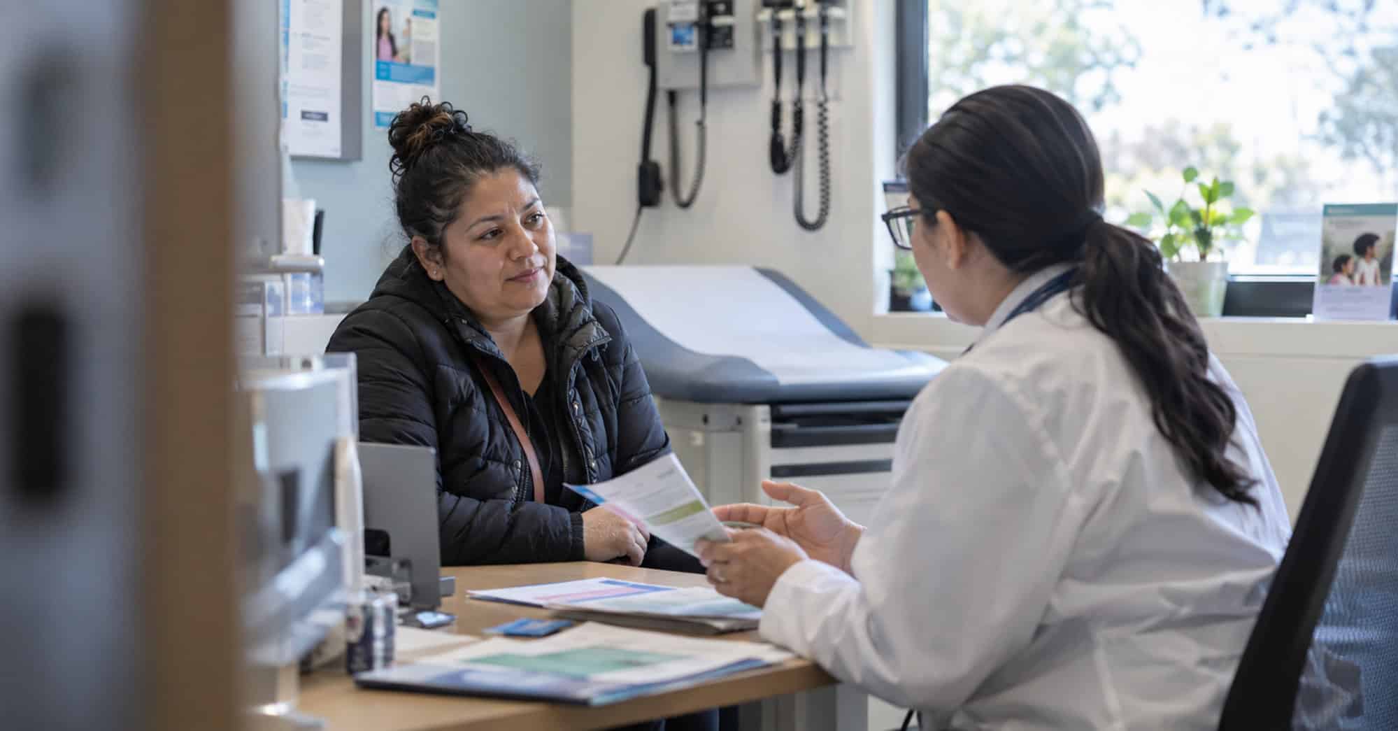 Inmigrante adulta conversa con una profesional sanitaria sobre atención médica y seguro de salud en una clínica de Estados Unidos.
