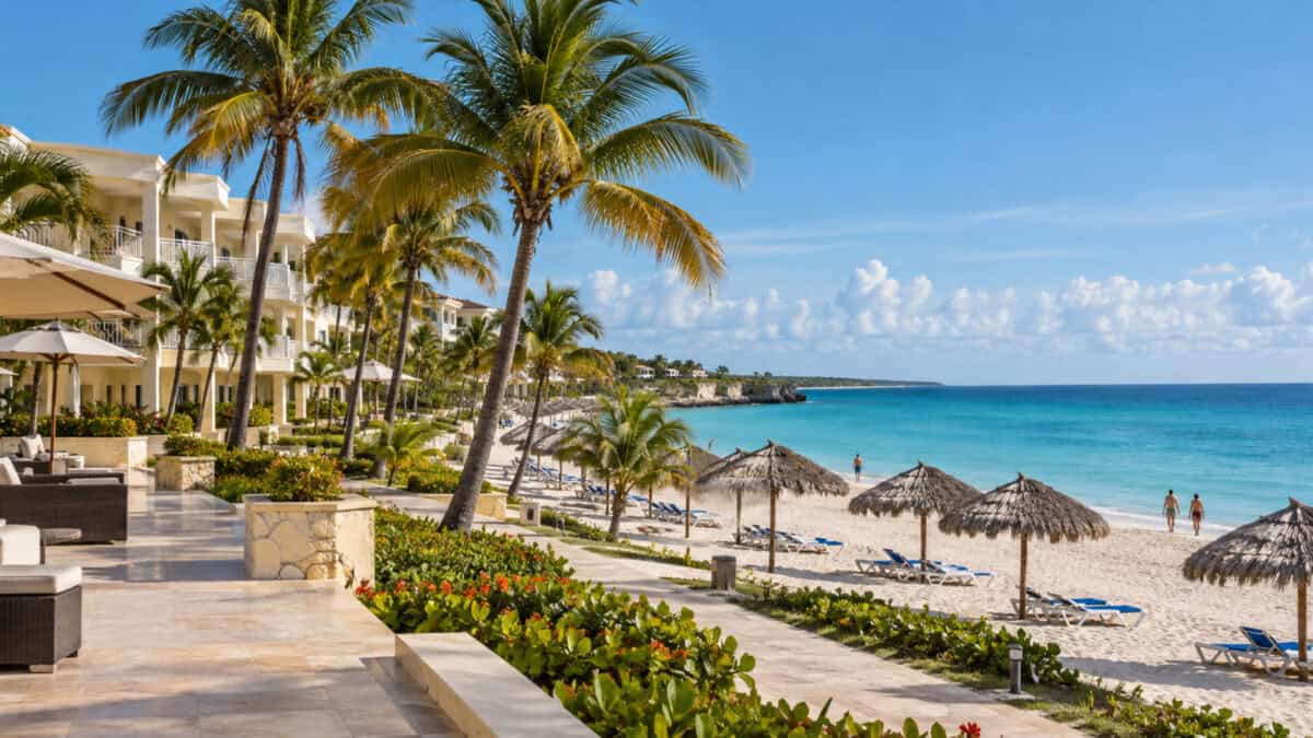 Hotel de arquitectura caribeña junto a una playa de arena clara, con palmeras, tumbonas vacías y pocos visitantes.