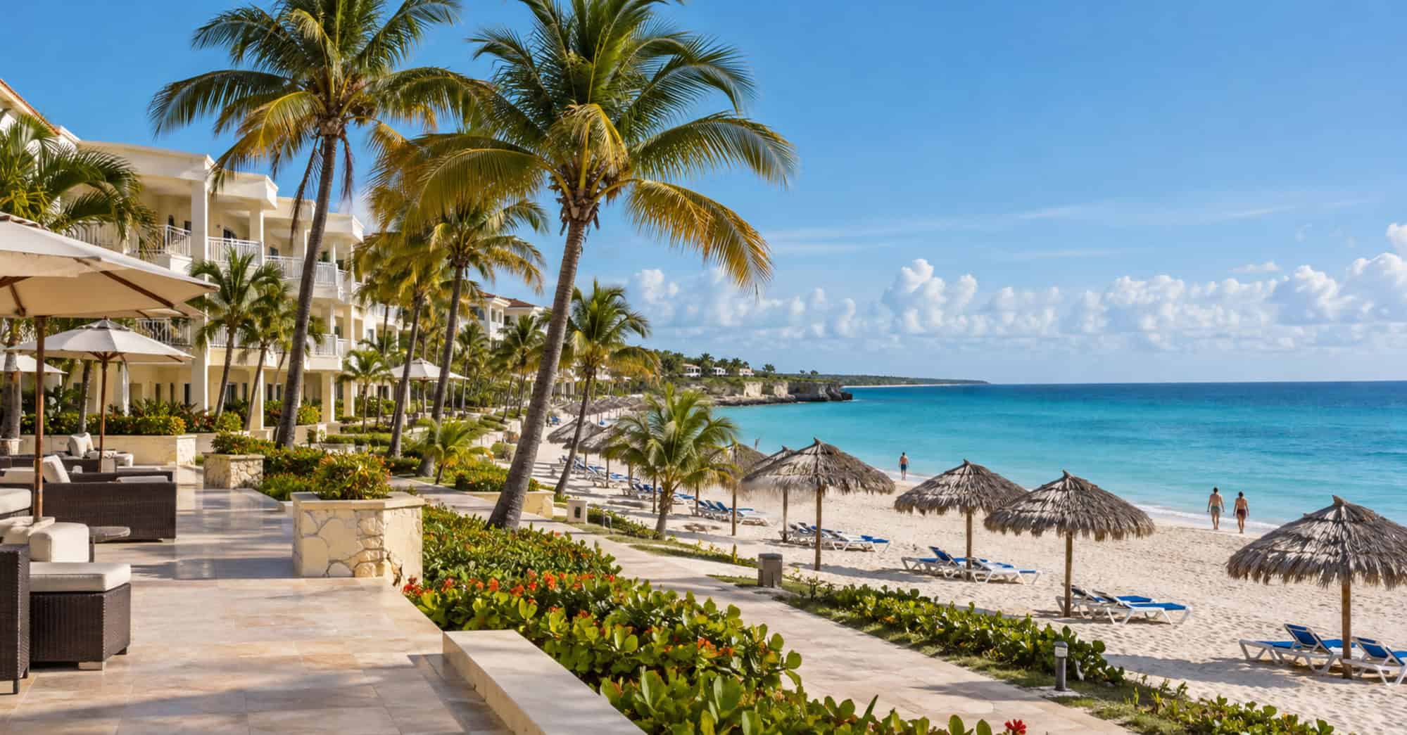 Hotel de arquitectura caribeña junto a una playa de arena clara, con palmeras, tumbonas vacías y pocos visitantes.