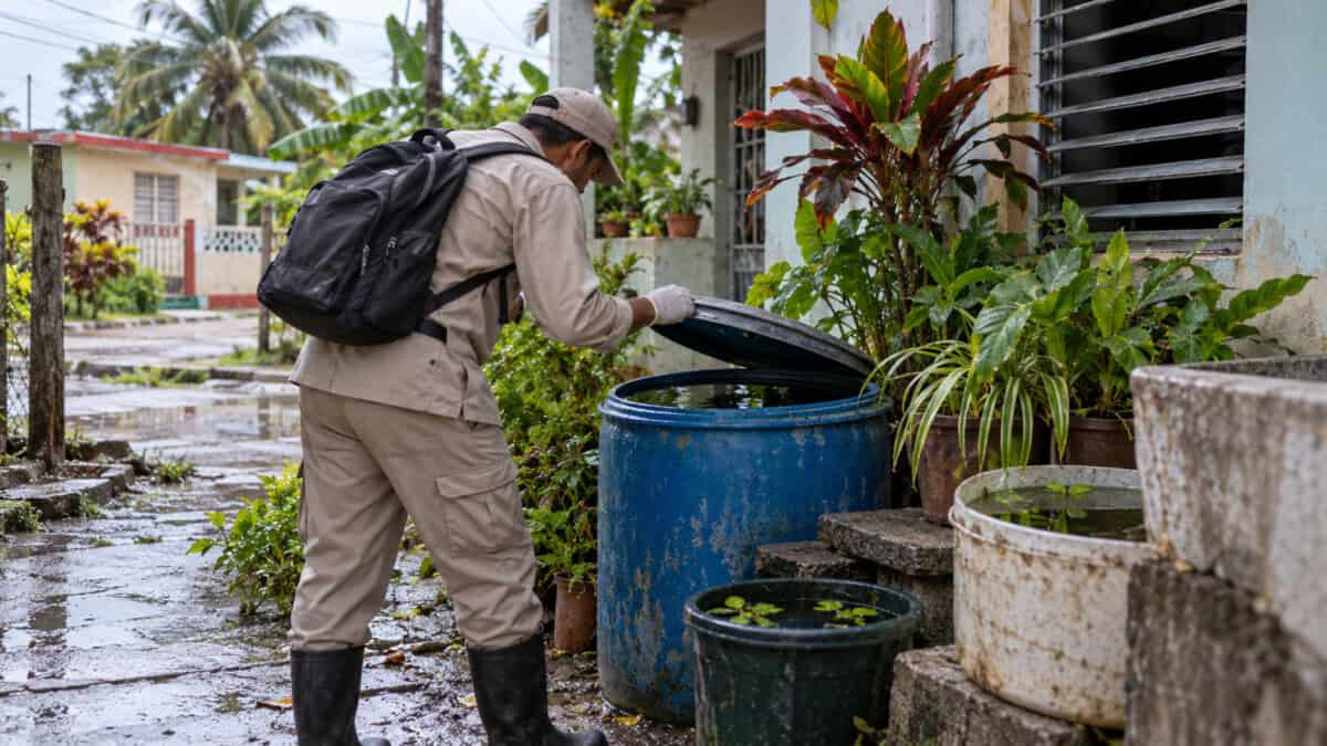 Trabajador sanitario inspecciona recipientes con agua acumulada junto a una vivienda en un barrio cubano.