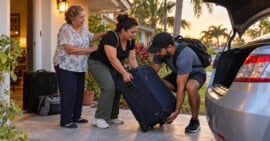 Una mujer y su hijo levantan una maleta junto a un automóvil en Miami mientras una familiar mayor los despide