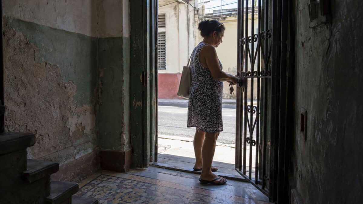 Mujer abre la reja de una vivienda cubana, vista desde un zaguán con paredes desgastadas y piso de mosaico.