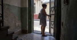 Mujer abre la reja de una vivienda cubana, vista desde un zaguán con paredes desgastadas y piso de mosaico.