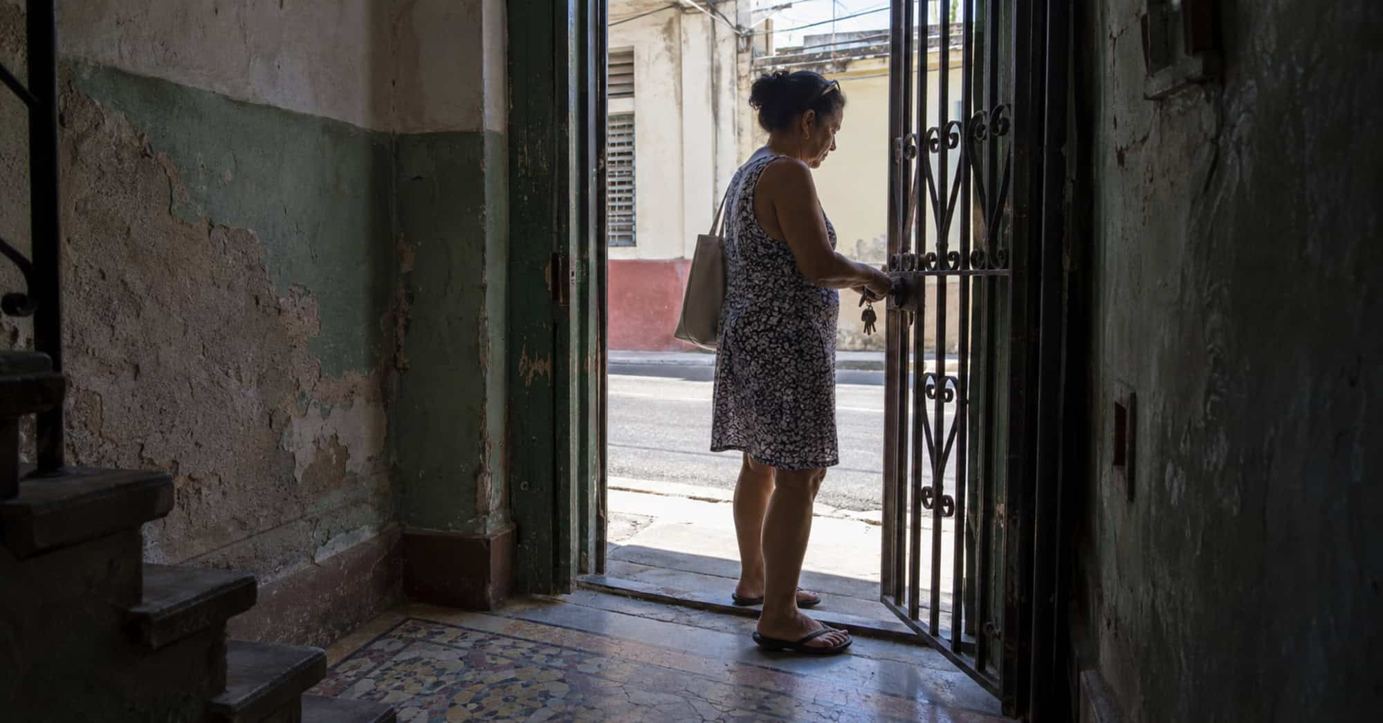 Mujer abre la reja de una vivienda cubana, vista desde un zaguán con paredes desgastadas y piso de mosaico.